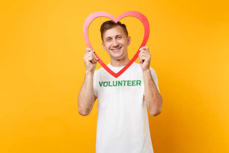 Portrait of happy man in white t-shirt with written inscription green title volunteer hold red wooden heart isolated on yellow background. Voluntary free assistance help charity grace work conceptの写真素材
