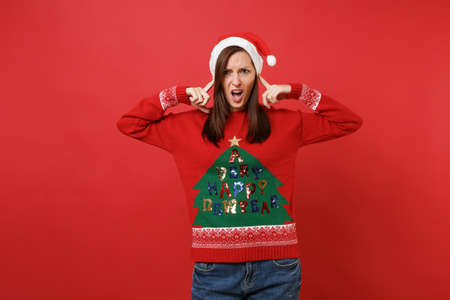 Dissatisfied young Santa girl in knitted sweater, Christmas hat covering ears with fingers isolated on bright red background. Happy New Year 2019 celebration holiday party concept. Mock up copy spaceの写真素材