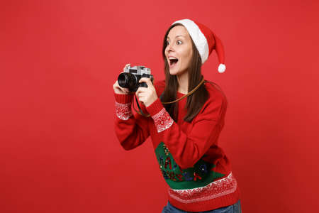 Surprised young Santa girl taking pictures on retro vintage photo camera, keeping mouth wide open isolated on red background. Happy New Year 2019 celebration holiday party concept. Mock up copy spaceの写真素材