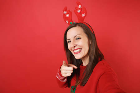 Close up selfie shot of smiling Santa girl in fun decorative deer horns pointing finger on camera isolated on red background. Happy New Year 2019 celebration holiday party concept. Mock up copy spaceの写真素材
