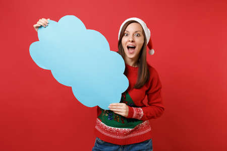 Surprised young Santa girl keeping mouth wide open hold blue empty blank Say cloud, speech bubble isolated on red background. Happy New Year 2019 celebration holiday party concept. Mock up copy spaceの写真素材