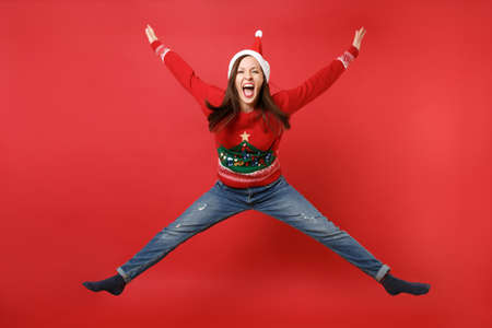 Crazy young Santa girl in Christmas hat jumping, spreading hands and legs, screaming isolated on bright red wall background. Happy New Year 2019 celebration holiday party concept. Mock up copy spaceの写真素材