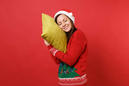 Relaxed young Santa girl in Christmas hat holding pillow keeping eyes closed sleeping isolated on bright red wall background. Happy New Year 2019 celebration holiday party concept. Mock up copy spaceの写真素材