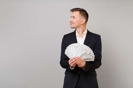 Handsome young business man in black suit looking aside holding lots bunch of dollars banknotes, cash money isolated on grey background. Achievement career wealth business concept. Mock up copy spaceの写真素材