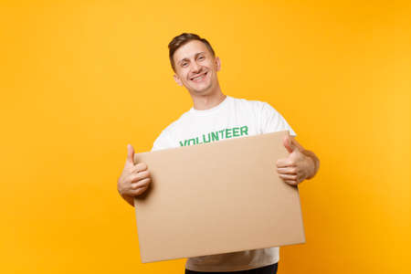 Portrait of good kind man in white t-shirt with written inscription green title volunteer with big cardboard box isolated on yellow background. Voluntary free assistance help, charity grace conceptの写真素材