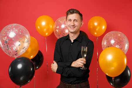 Handsome young man in black classic shirt holding glass of champagne, celebrating on red background air balloons. St. Valentine's, Women's Day, Happy New Year, birthday mockup holiday party conceptの写真素材