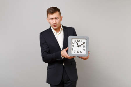 Portrait of puzzled young business man in classic black suit and shirt holding square clock isolated...