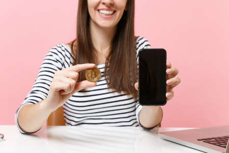 Cropped image of woman holding bitcoin metal coin of golden color future currency mobile phone with blank empty screen sit at desk isolated on pink background. Achievement business career. Copy spaceの写真素材