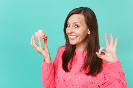 Smiling attractive young woman in knitted pink sweater showing OK gesture holding in hand cake isolated on blue turquoise wall background studio portrait. People lifestyle concept. Mock up copy spaceの写真素材