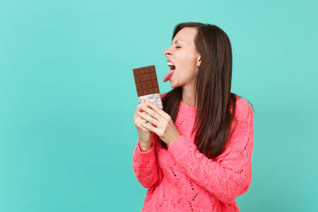 Funny young woman in knitted pink sweater keeping eyes closed, licking chocolate bar in hands isolated on blue turquoise wall background, studio portrait. People lifestyle concept. Mock up copy spaceの写真素材