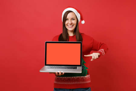 Cheerful young Santa girl pointing index finger on laptop pc computer with blank empty screen isolated on red background. Happy New Year 2019 celebration holiday party concept. Mock up copy spaceの写真素材