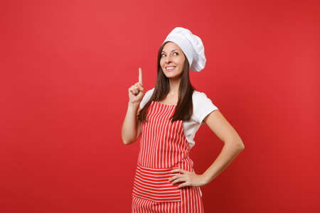 Housewife female chef cook or baker in striped apron, white t-shirt, toque chefs hat isolated on red wall background. Beautiful housekeeper woman pointing index finger up. Mock up copy space conceptの写真素材