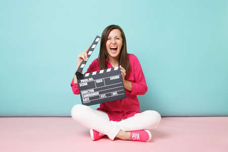 Full length shot woman in rose shirt white pants sitting on floor with film making clapperboard isolated on pink blue pastel wall background studio. Fashion lifestyle concept Mock up copy spaceの写真素材
