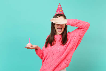 Young woman in knitted pink sweater, birthday hat keeping mouth wide open, covering eyes with hand, holding cake with candle isolated on blue background. People lifestyle concept. Mock up copy spaceの写真素材