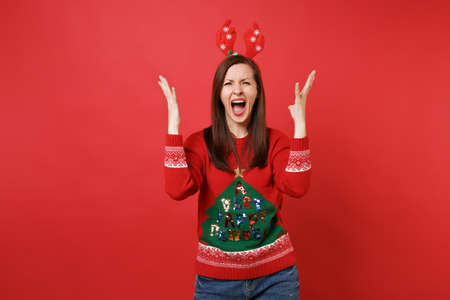 Angry young Santa girl in knitted sweater, fun decorative deer horns screaming, spreading hands isolated on red background. Happy New Year 2019 celebration holiday party concept. Mock up copy spaceの写真素材