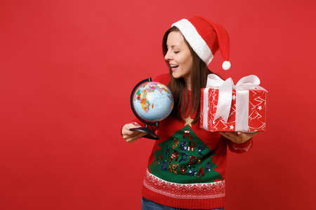 Pretty young Santa girl in Christmas hat looking on world globe holding red box with gift present isolated on red background. Happy New Year 2019 celebration holiday party concept. Mock up copy spaceの写真素材