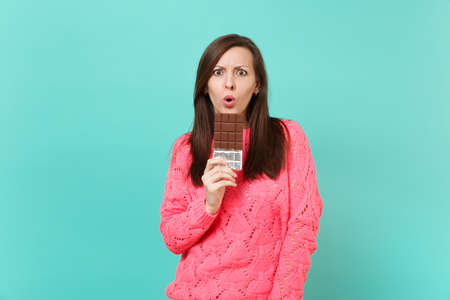 Shocked young girl in knitted pink sweater hold in hand chocolate bar keeping mouth wide open isolated on blue turquoise wall background, studio portrait. People lifestyle concept. Mock up copy spaceの写真素材
