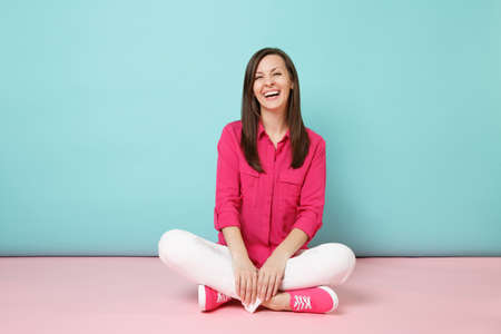 Full length portrait of smiling young woman in rose shirt blouse, white pants sitting on floor isolated on bright pink blue pastel wall background studio Fashion lifestyle concept. Mock up copy spaceの写真素材