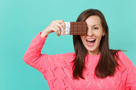 Cheerful young woman in knitted pink sweater holding in hand, covering eye with chocolate bar...