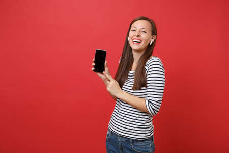 Laughing young woman with wireless earphones hold mobile phone with blank black empty screen listening music isolated on red background. People sincere emotions, lifestyle concept. Mock up copy spaceの写真素材