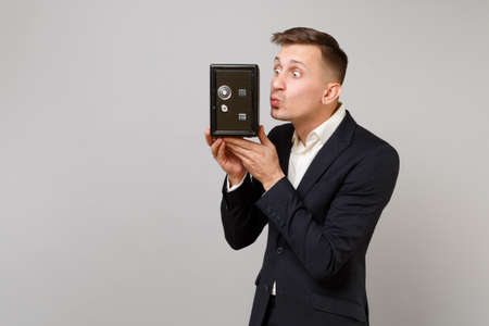 Funny young business man in suit blowing sending air kiss to metal bank safe for money accumulation in hand isolated on grey background. Achievement career wealth business concept. Mock up copy spaceの写真素材