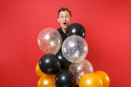 Amazed young man in black classic shirt holding air balloons, celebrating isolated on bright red...