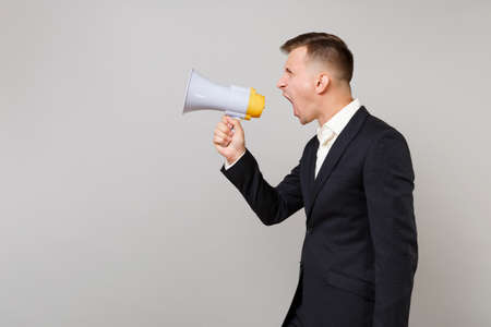 Side view of young business man in classic black suit and white shirt screaming on megaphone isolated on grey wall background in studio. Achievement career wealth business concept. Mock up copy spaceの写真素材