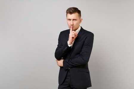 Serious young business man in classic black suit shirt say hush be quiet with finger on lips, shhh gesture isolated on grey background. Achievement career wealth business concept. Mock up copy space