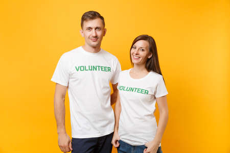 Portrait of young colleagues couple in white t-shirt with written inscription green title volunteer isolated on yellow background. Voluntary free work, assistance help, charity grace teamwork conceptの写真素材
