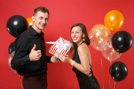 Young couple in black clothes hold gift box celebrating birthday holiday party isolated on bright red background air balloons St. Valentine International Women Day Happy New Year 2019 concept.の写真素材