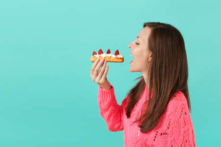 Side view of young woman in knitted pink sweater with closed eyes hold in hand, eating eclair cake...