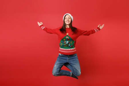 Relaxed young Santa girl with closed eyed jumping, meditate spreading hands, keeping legs crossed isolated on red background. Happy New Year 2019 celebration holiday party concept. Mock up copy spaceの写真素材