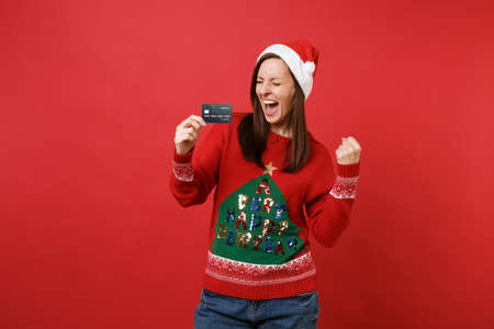 Happy young Santa girl with closed eyes screaming clenching fist like winner, holding credit card isolated on red background. Happy New Year 2019 celebration holiday party concept. Mock up copy spaceの写真素材