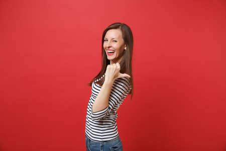 Portrait of cheerful laughing young woman in striped clothes pointing thumb behind her back isolated on bright red wall background. People sincere emotions, lifestyle concept. Mock up copy spaceの写真素材