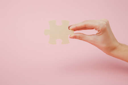 Close up cropped photo of hand holding wooden jigsaw puzzle piece isolated on pastel pink wall...
