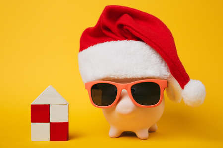 Pink piggy money bank with sunglasses Christmas hat, toy wooden house isolated on yellow wall background. Money accumulation investment banking services wealth concept. Copy space advertising mock upの写真素材