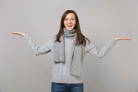 Joyful young woman in gray sweater, scarf spreading, pointing hands aside isolated on grey wall background. Healthy fashion lifestyle, people sincere emotions, cold season concept. Mock up copy spaceの写真素材