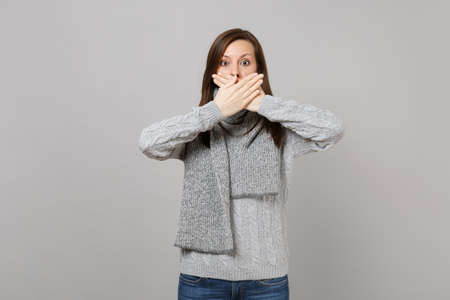 Young woman in gray sweater, scarf covering mouth with hands isolated on grey background, studio portrait. Healthy fashion lifestyle, people sincere emotions, cold season concept. Mock up copy spaceの写真素材
