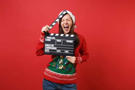 Crazy young Santa girl with closed eyes screaming, holding classic black film making clapperboard isolated on red background. Happy New Year 2019 celebration holiday party concept. Mock up copy spaceの写真素材