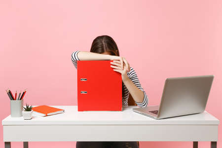 Tired woman leaning on red folder with paper documents and working on project while sitting at office with laptop isolated on pastel pink background. Achievement business career concept. Copy spaceの写真素材