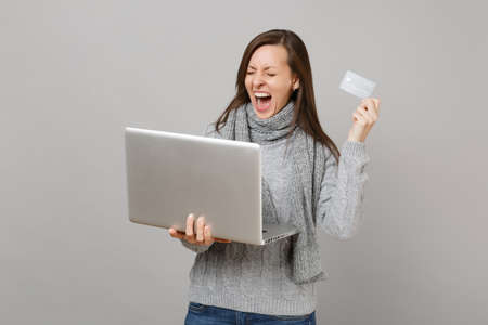 Screaming woman in sweater, scarf with closed eyes work on laptop pc computer holding credit bank...