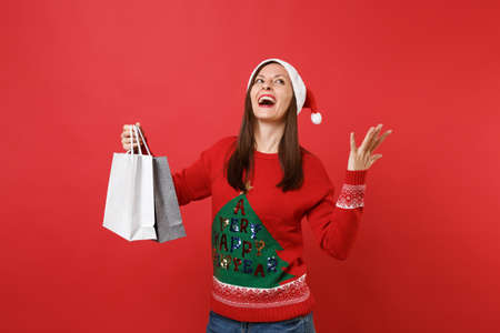 Happy Santa girl looking up, spreading hands, holding packages bags with purchases after shopping isolated on red background. Happy New Year 2019 celebration holiday party concept. Mock up copy spaceの写真素材
