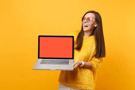 Laughing young woman in heart eyeglasses holding laptop pc computer with blank black empty screen isolated on bright yellow background. People sincere emotions, lifestyle concept. Advertising areaの写真素材