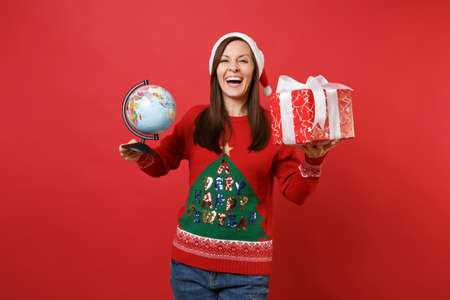 Laughing young Santa girl in Christmas hat holding world globe, red box with gift present isolated on bright red background. Happy New Year 2019 celebration holiday party concept. Mock up copy spaceの写真素材