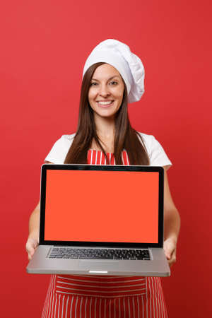 Housewife female chef cook or baker in striped apron white t-shirt toque chefs hat isolated on red wall background. Woman hold laptop blank screen for promotional content Mock up copy space conceptの写真素材