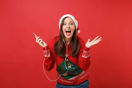 Excited young Santa girl with earphones keeping mouth wide open, spreading hands holding mobile phone listening music isolated on red background. Happy New Year 2019 celebration holiday party conceptの写真素材