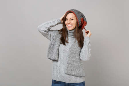 Pretty young woman in gray sweater hat and scarf putting hands on head isolated on grey background...