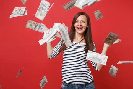 Smiling young woman in striped clothes spreading hands, scattering lots of dollars, standing under...