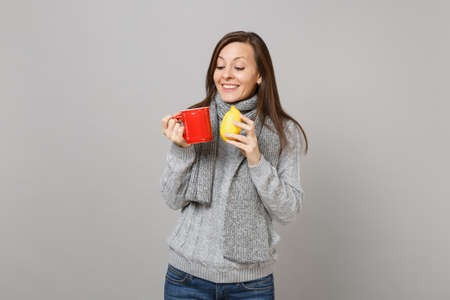 Pretty young woman in gray sweater, scarf holding lemon, red cup of tea isolated on grey wall background. Healthy fashion lifestyle, people sincere emotions, cold season concept. Mock up copy spaceの写真素材