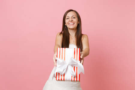 Portrait of smiling bride woman in lace white wedding dress showing red box with gift present on camera isolated on pink pastel background. Wedding celebration concept. Copy space for advertisementの写真素材